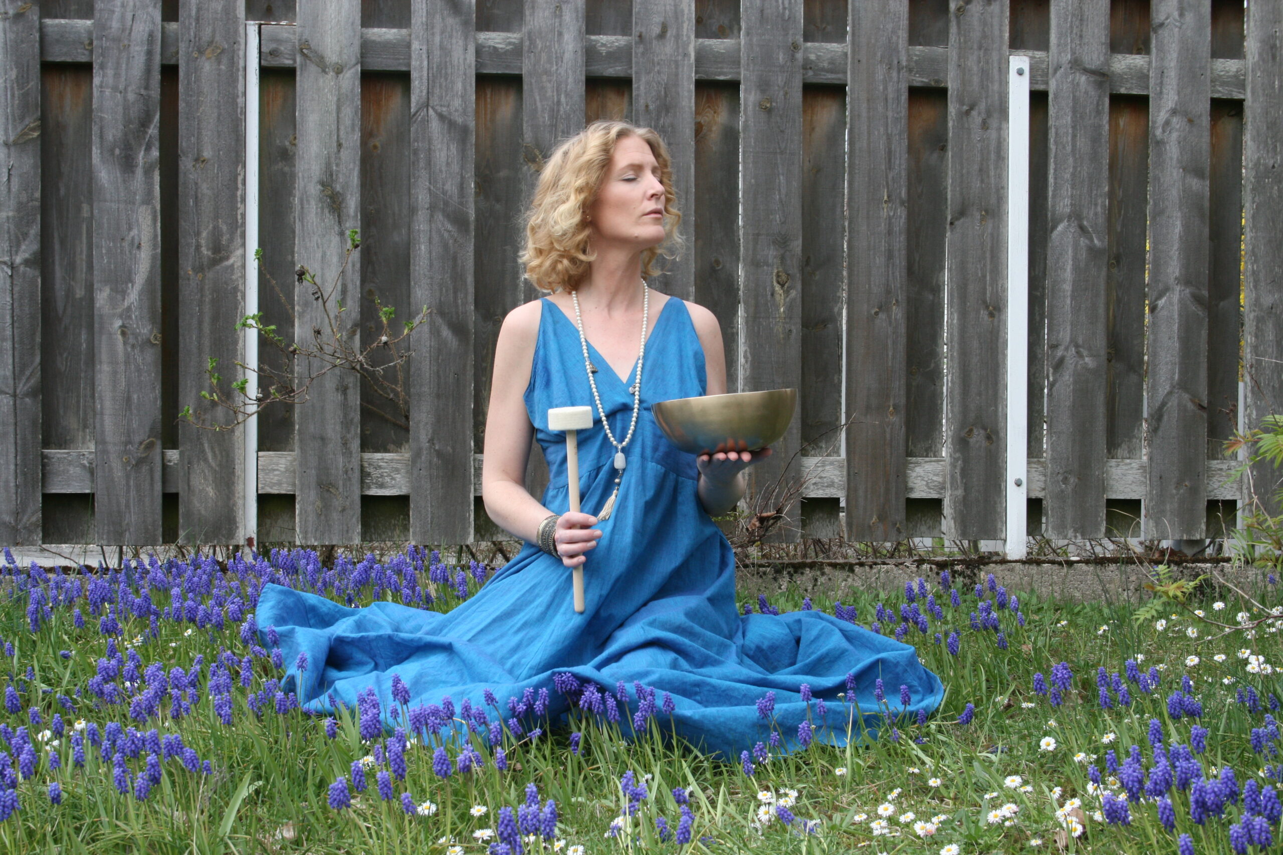 Klangskålsmeditation, kvinna i blå klänning håller i klangskål, sitter utomhus på gräset som är fullt med blå blommor